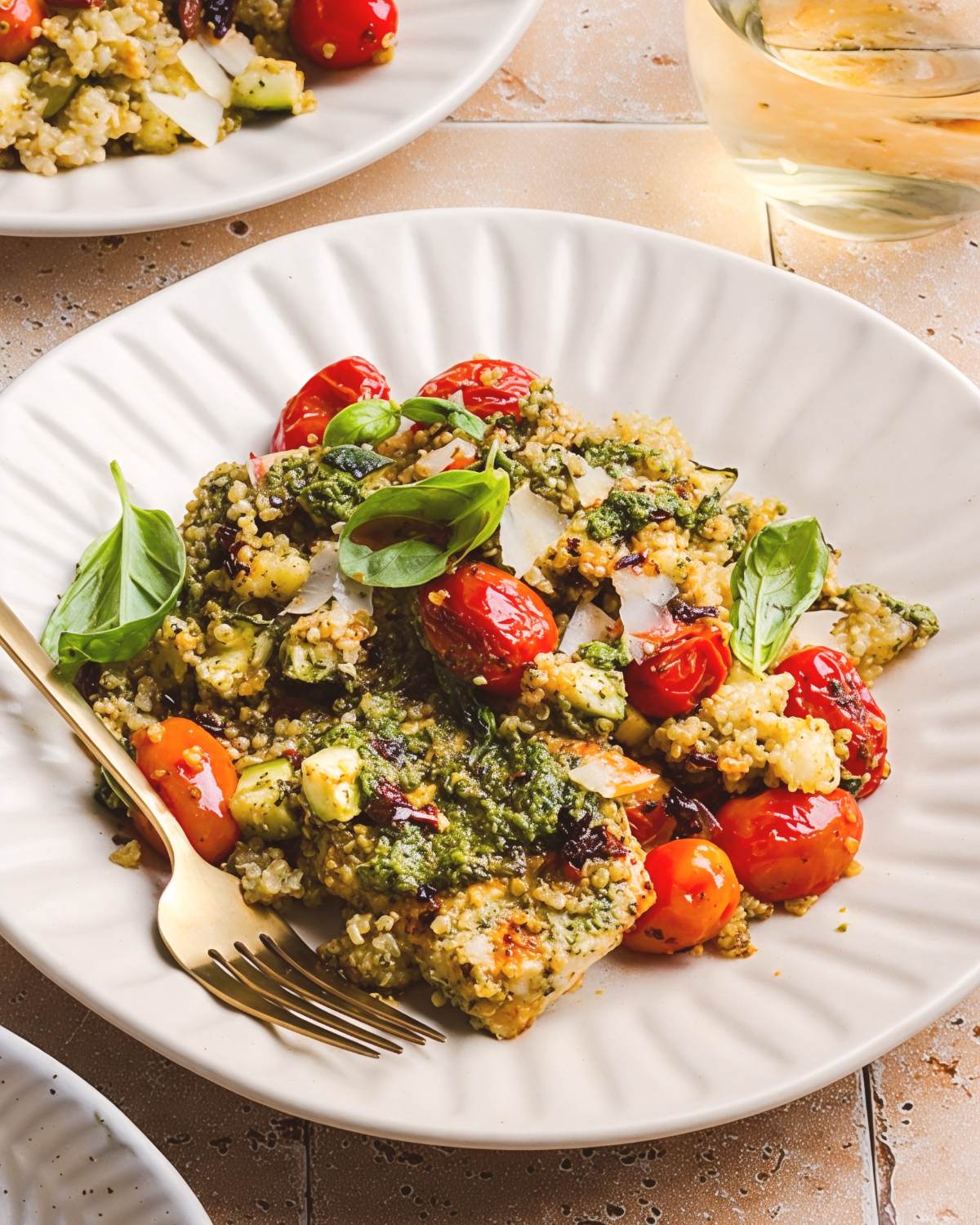 A plate of healthy pesto chicken and quinoa bake topped with blistered cherry tomatoes, shaved parmesan, and fresh basil, served with a gold fork as a nutritious twist on a classic dump and bake pasta dish