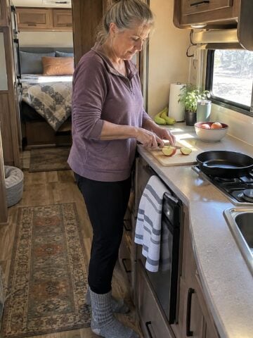 Early morning in an RV galley kitchen, a woman in her fifties stands alone at the narrow counter, cutting an apple beside a small skillet.