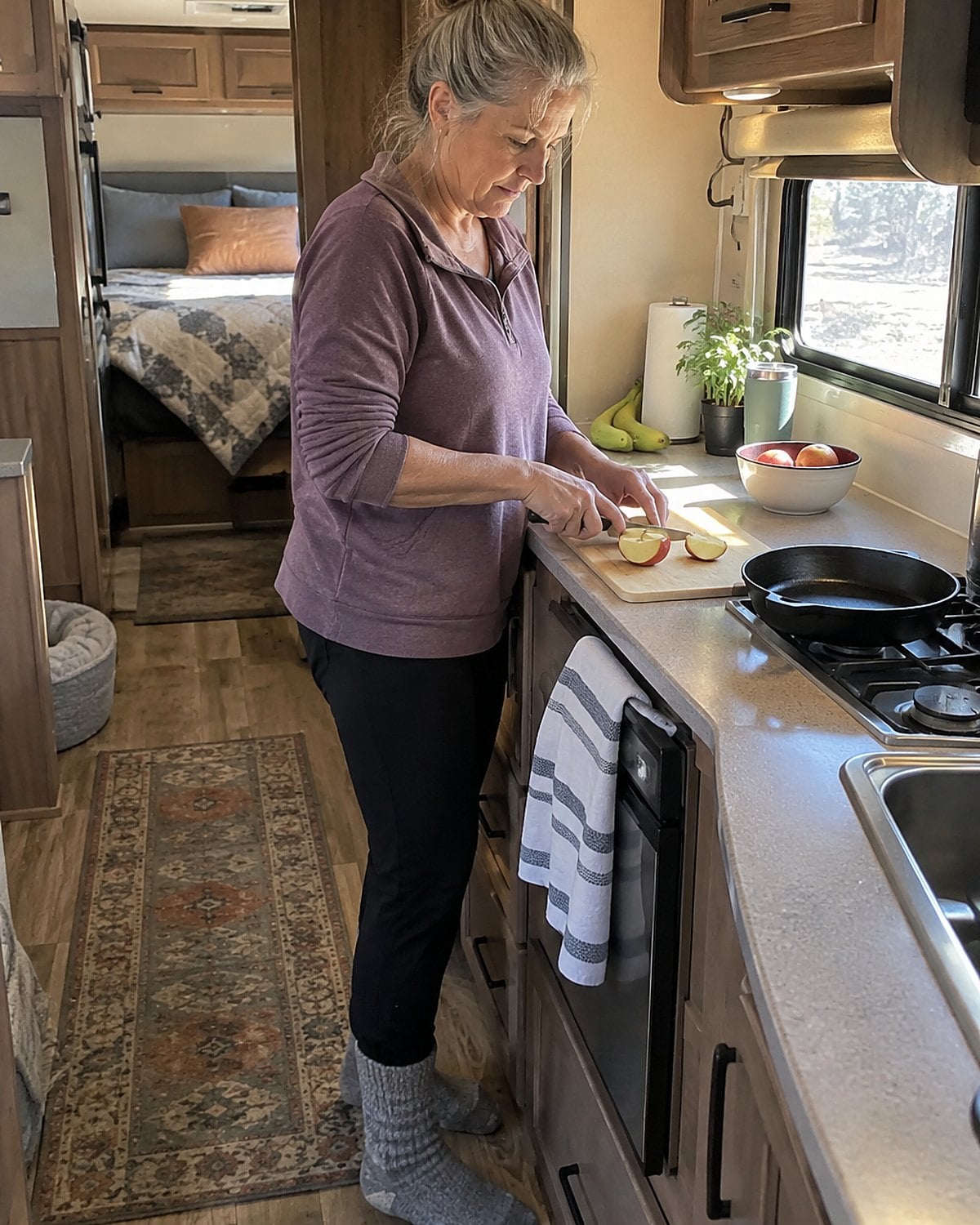 Early morning in an RV galley kitchen, a woman in her fifties stands alone at the narrow counter, cutting an apple beside a small skillet.