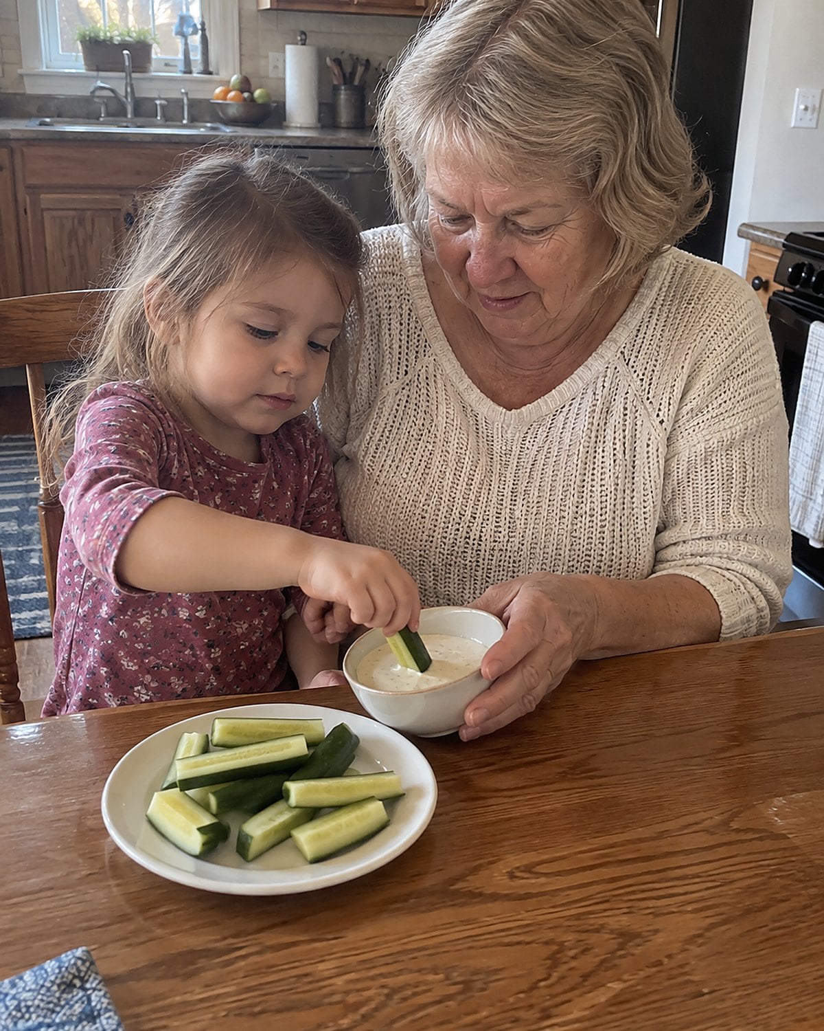 A grandchild's hand dipping a short cucumber stick into a little bowl of ranch.