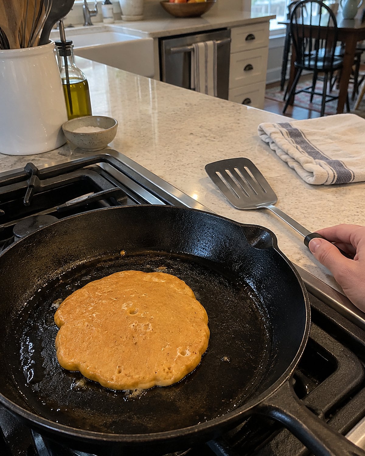 A small pumpkin pancake cooking in a skillet while a spatula waits beside it.