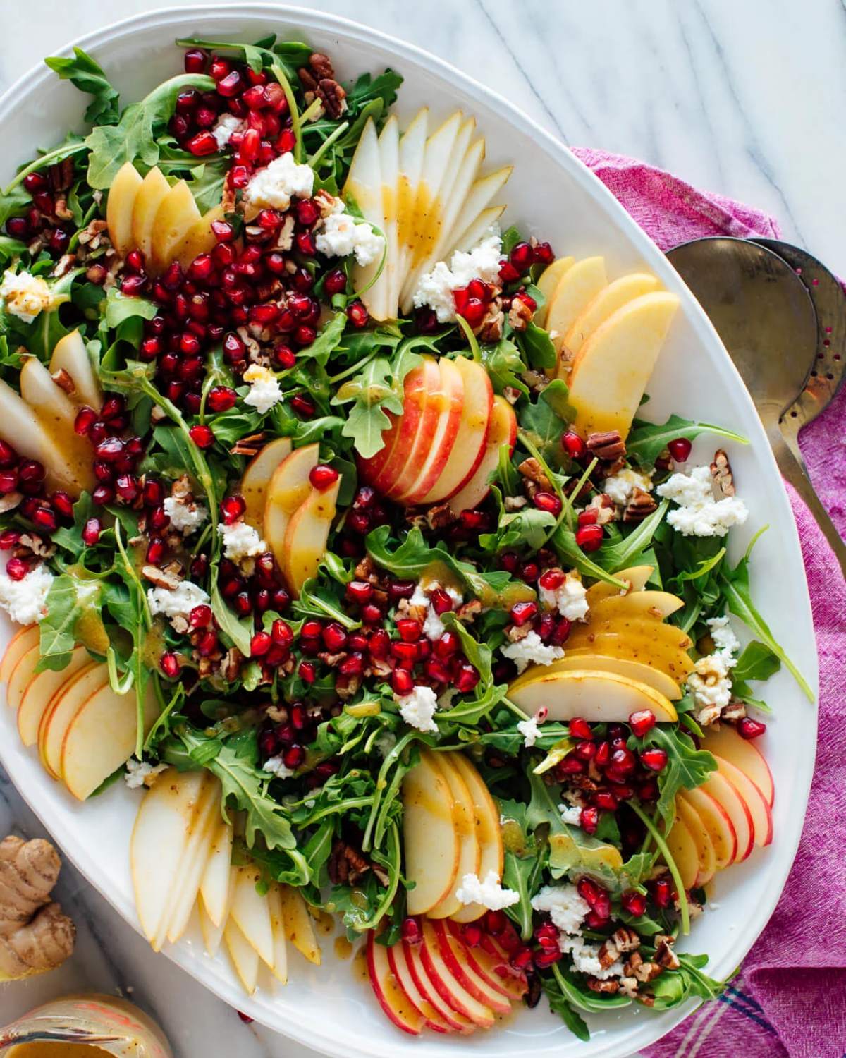 A large oval serving platter filled with a fresh green salad topped with sliced pears, apples, juicy pomegranate seeds, goat cheese, and ginger dressing for Thanksgiving vegetable side dishes.