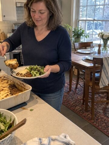 A woman in her fifties sits alone at a dining table set for two around noon, reaching for a spoonful of casserole from a serving dish.