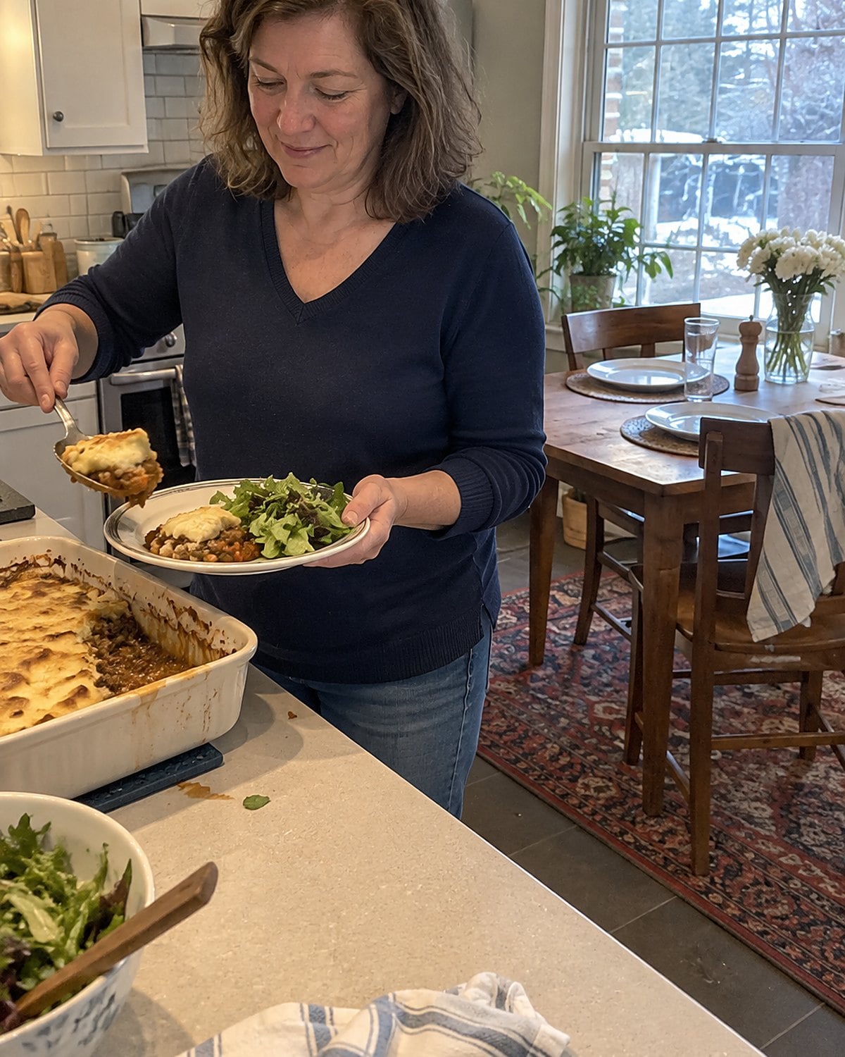 A woman in her fifties sits alone at a dining table set for two around noon, reaching for a spoonful of casserole from a serving dish.