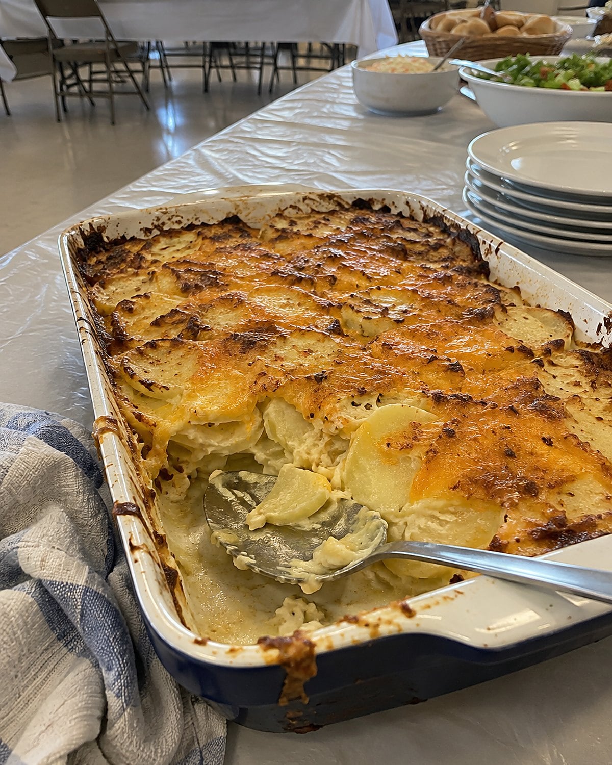 A baking dish of cheesy potatoes on a church hall table, its browned corner partly served.