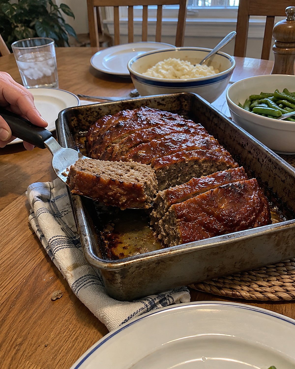 A sliced meatloaf in its pan on a family dining table, with a broad spatula under one slice.