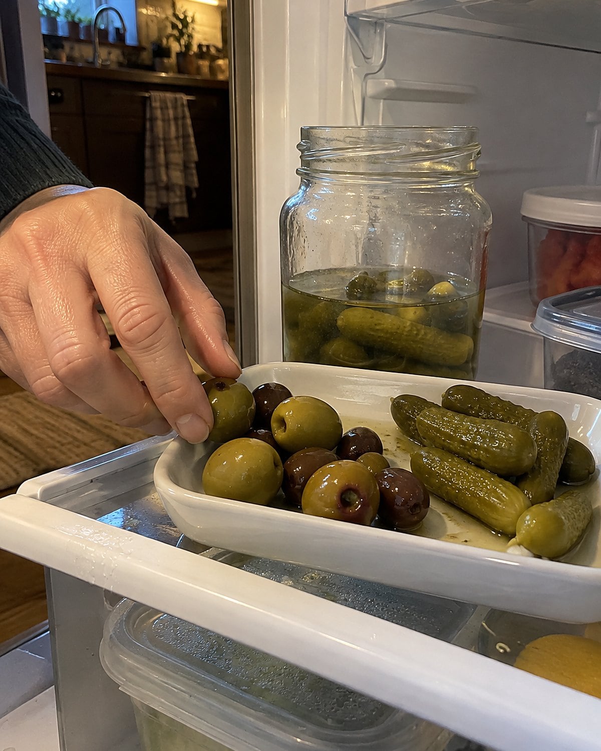 A small tray of pickles and olives on a refrigerator shelf, with an open jar behind it.