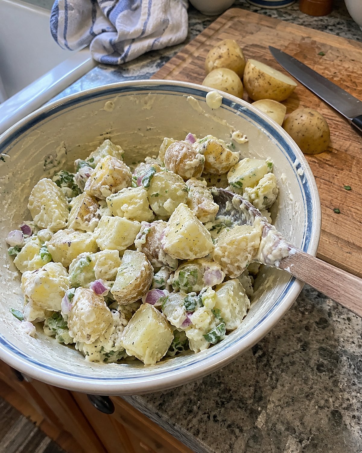 A bowl of potato salad beside a thin knife and a few cooked potatoes on a cutting board.