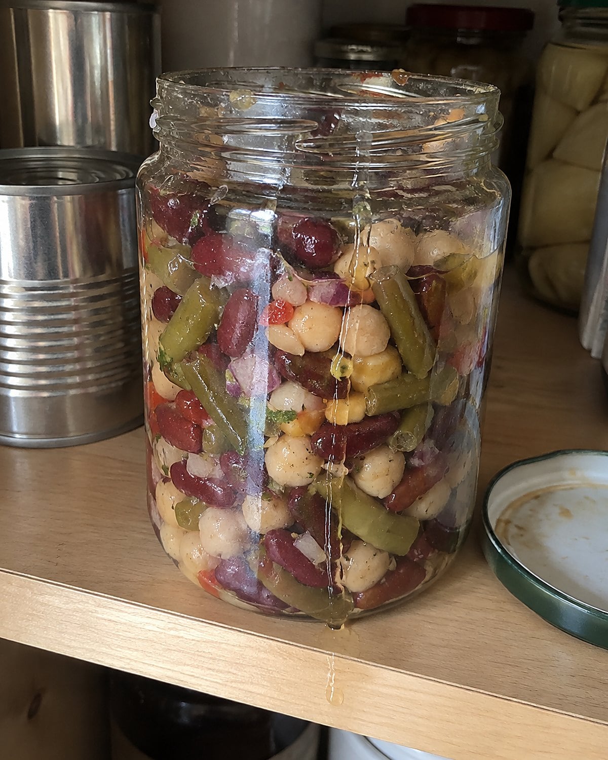 Three bean salad in a plain glass jar on a pantry shelf beside unopened cans.