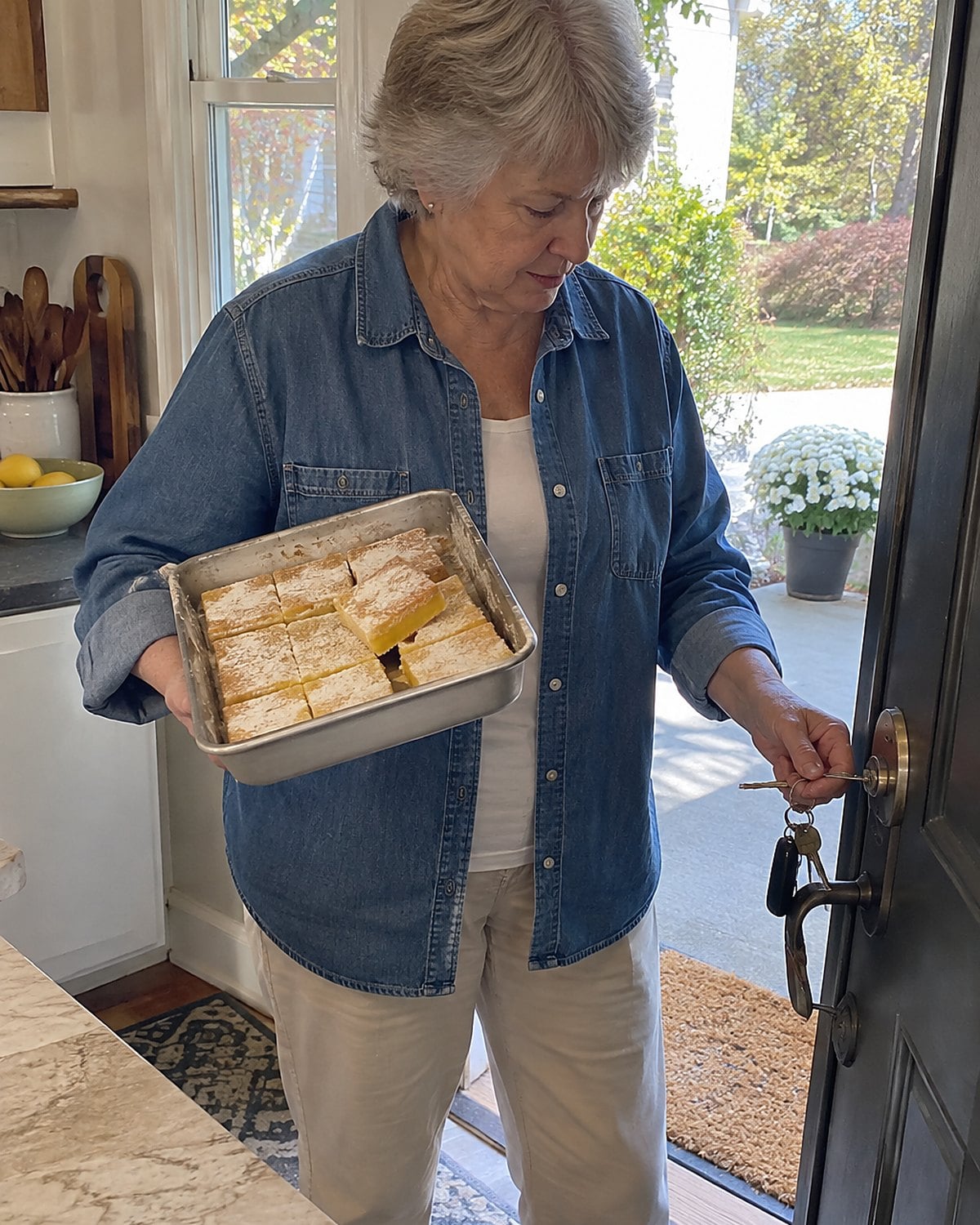 Hands holding a pan of lemon bars at a porch door in daylight, with keys visible in one hand.