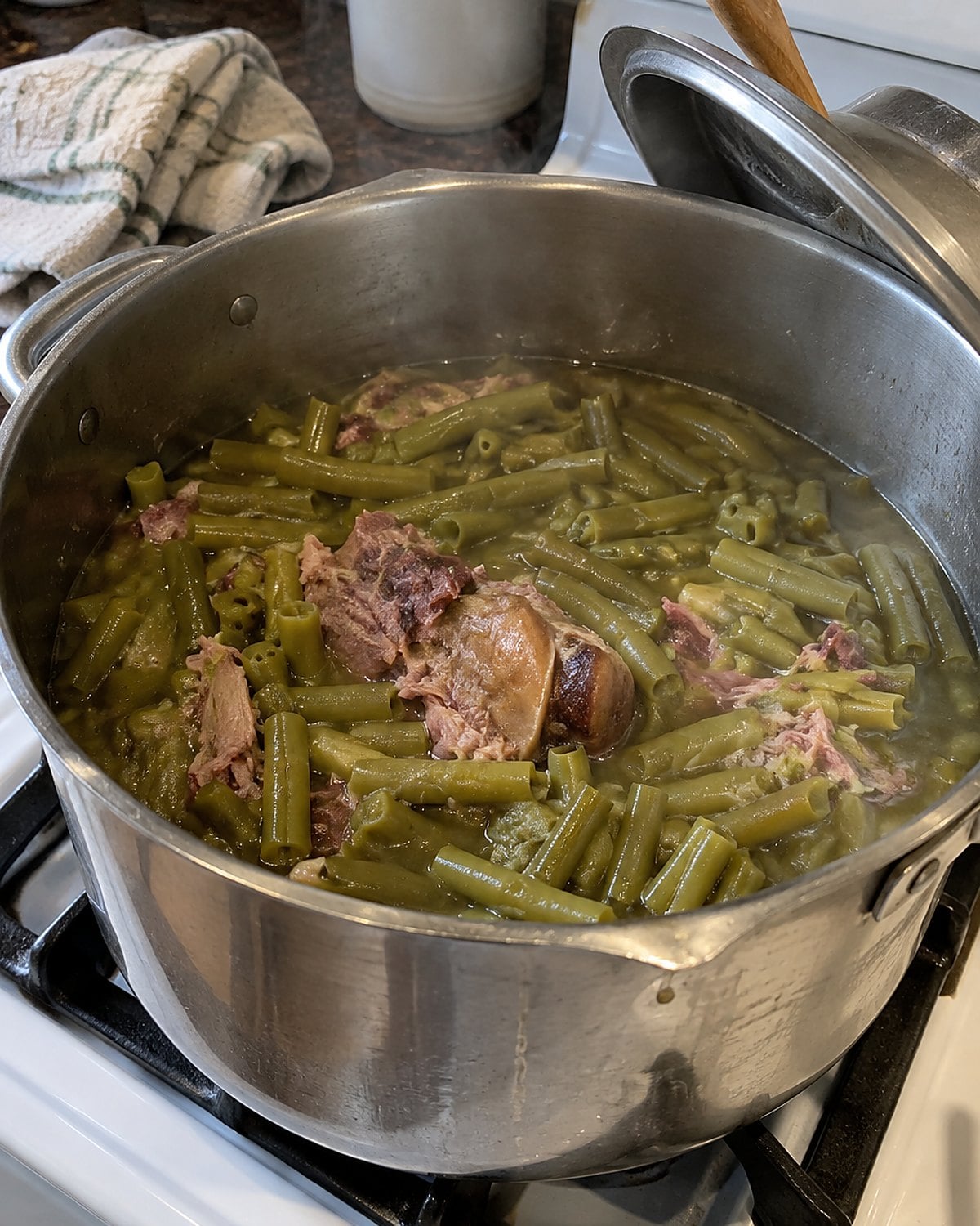 A dented pot of green beans and ham hock on a white stove, its lid propped on a wooden spoon.