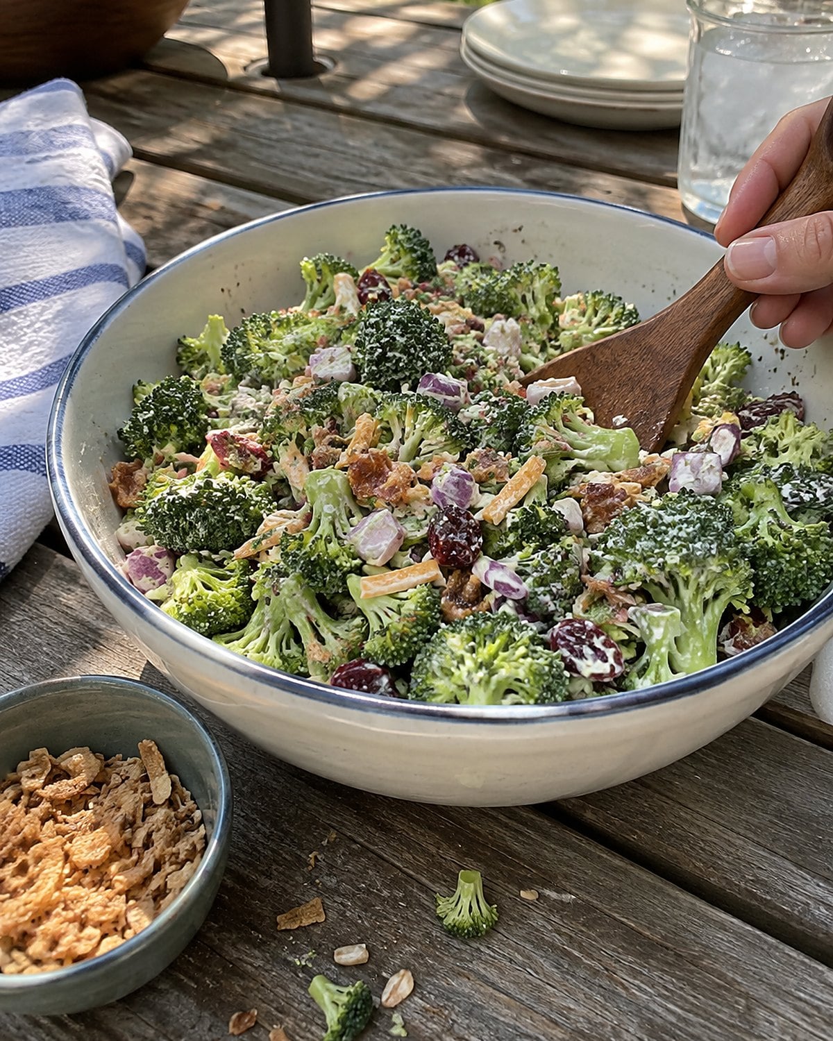 A bowl of chopped broccoli salad on a picnic table, with a small cup of crunchy topping beside it.
