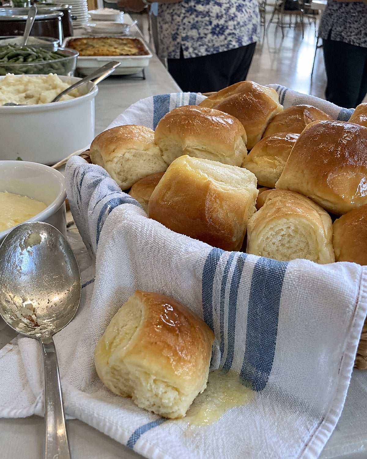 A towel-lined basket of buttered dinner rolls at the end of a church hall serving table.