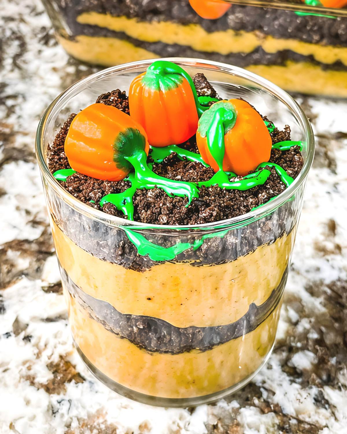 A clear glass filled with layered pumpkin spice pudding and crushed chocolate cookies, decorated on top with green icing vines and mini candy pumpkins.