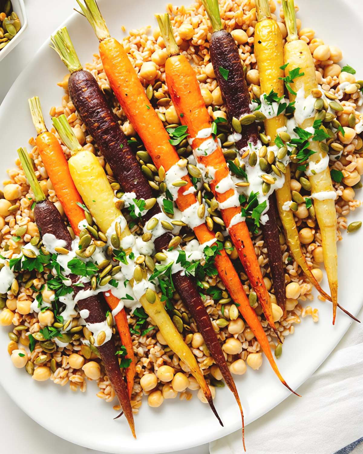 Rainbow roasted carrots served over a bed of farro and chickpeas, drizzled with herbed crรจme fraรฎche and pepitas for vibrant Thanksgiving vegetable side dishes.