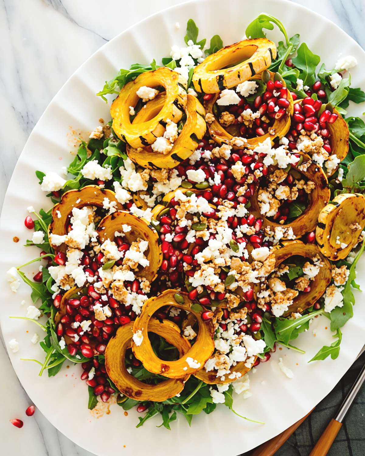 A large white platter featuring a roasted delicata squash and arugula salad topped with vibrant pomegranate seeds and crumbled cheese for Thanksgiving vegetable side dishes.