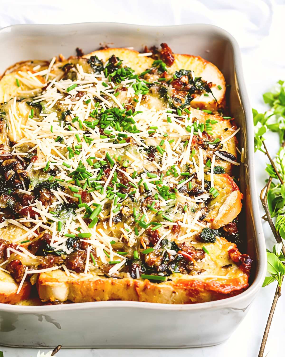 A baking dish filled with a savory sausage, kale, and mushroom strata topped with melted cheese and fresh chives as Thanksgiving breakfast ideas.