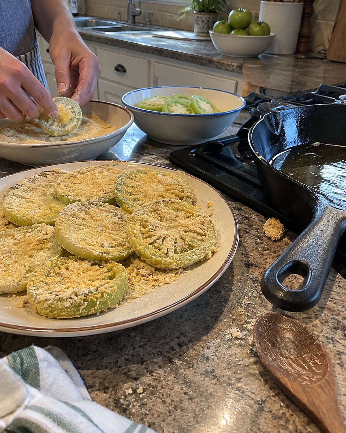 Coated green tomato slices wait beside a skillet on a flour-dusted counter.