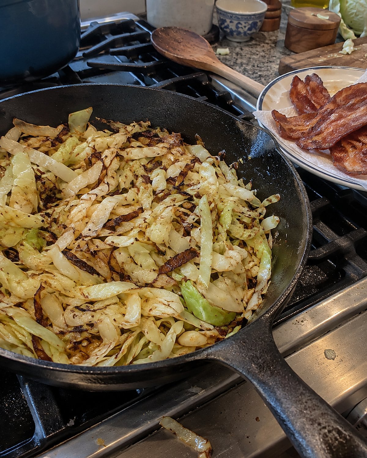 Cabbage ribbons brown in a skillet beside a plate holding cooked bacon.