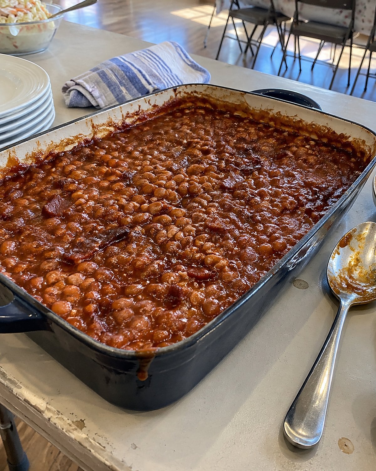 A baking dish of beans with a dark rim sits on a church hall table beside serving spoons.