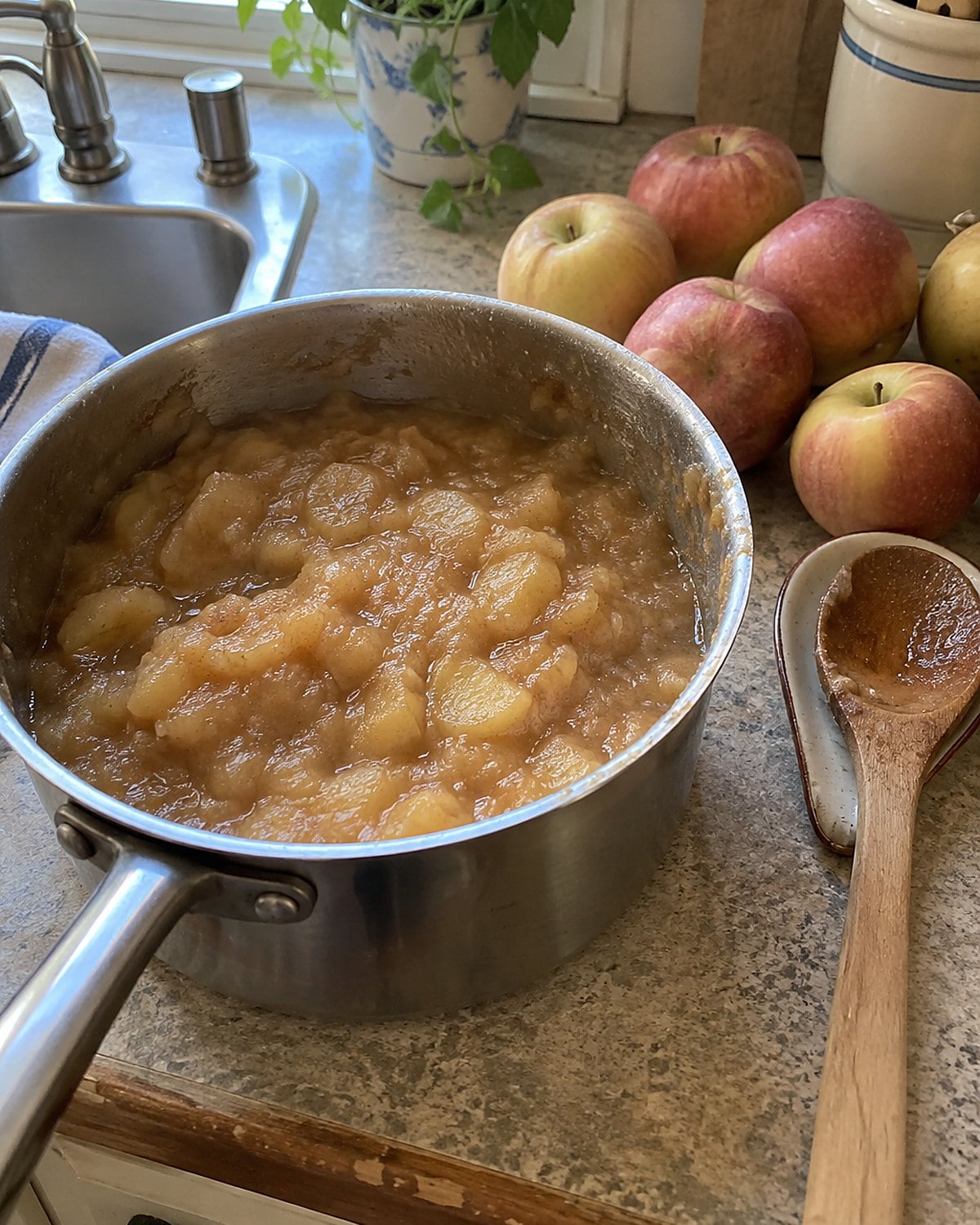 A saucepan of chunky applesauce beside several soft apples on a worn kitchen counter.