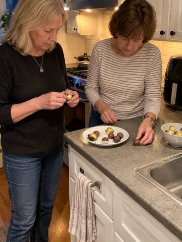 Two women friends at the counter beside an air fryer, unwrapping candy in the evening by the stove light, neither posing.