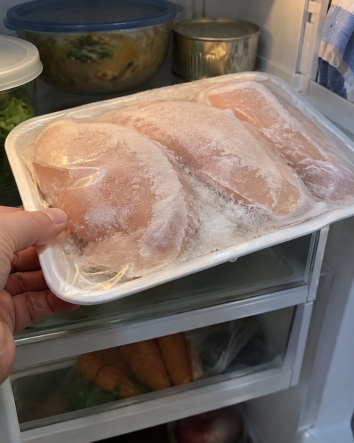 A frosty package of chicken breasts on a tray on a refrigerator shelf.