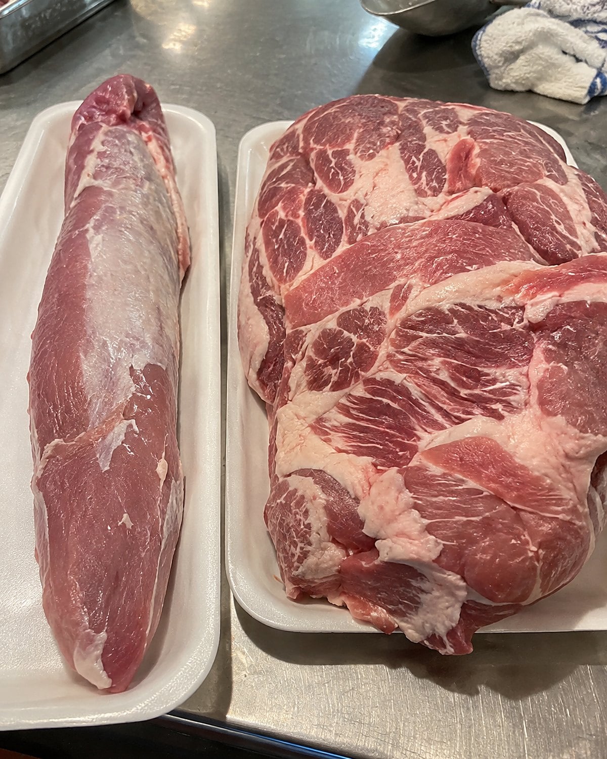 A pork tenderloin and a pork shoulder side by side on separate trays at a market meat counter.