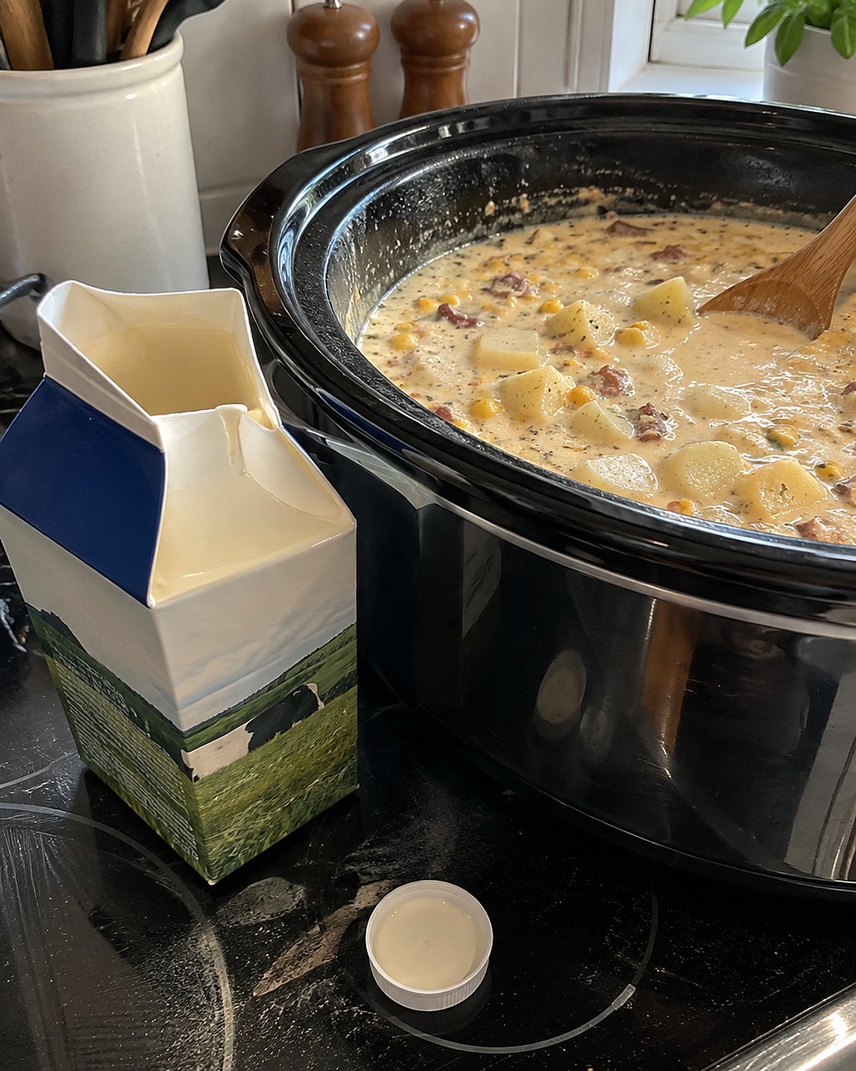 An open carton of cream beside a slow cooker of chowder on the stove.