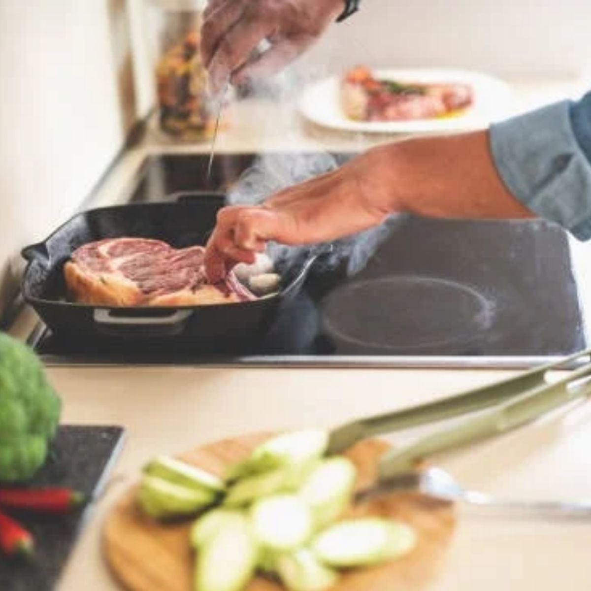 A cast-iron skillet smokes on a stove while a hand reaches to turn the meat