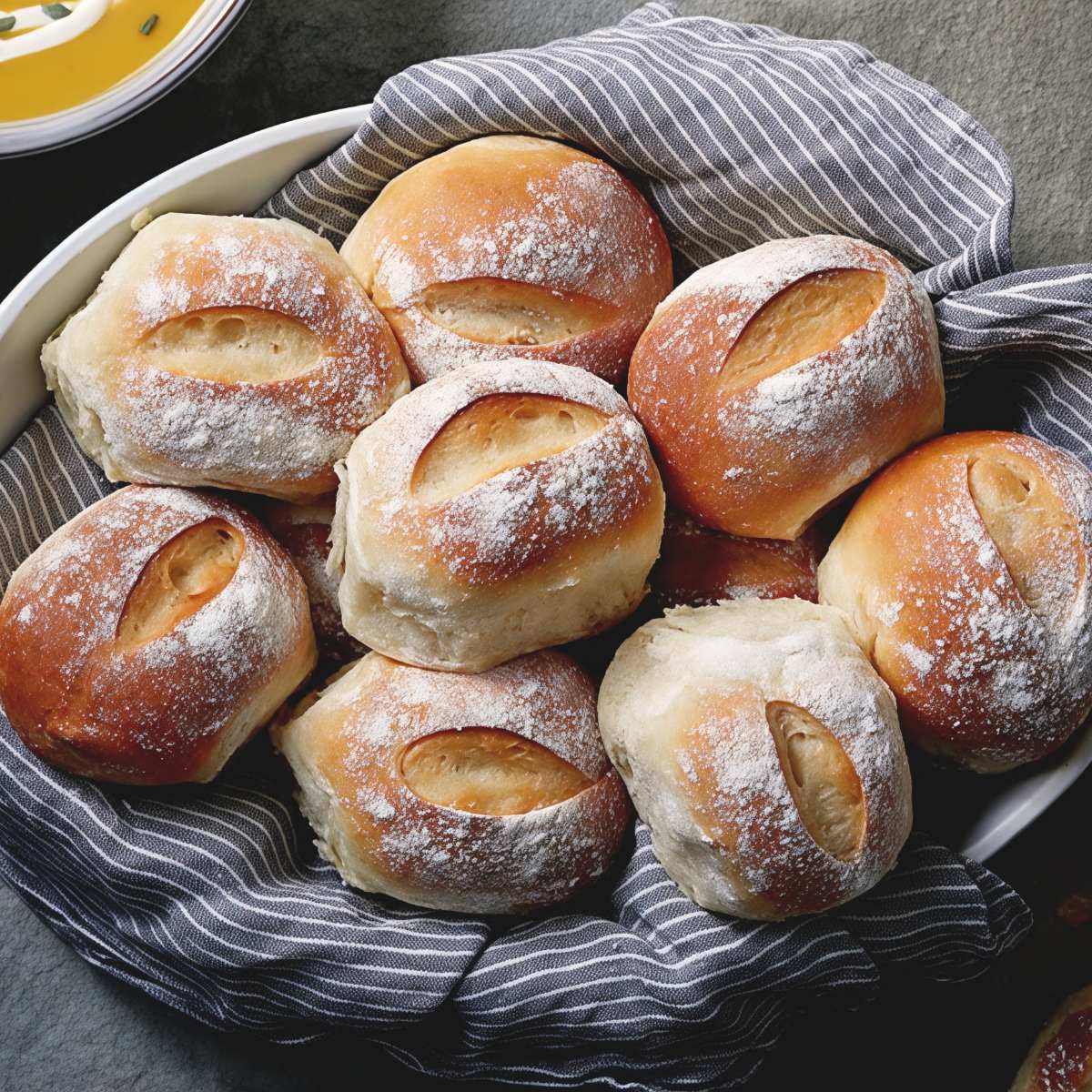 Freshly baked, golden-brown dinner rolls lightly dusted with flour and scored on top are nestled in a bowl lined with a striped cloth towel.