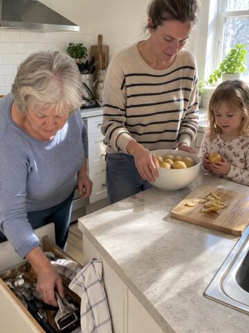 At a kitchen drawer pulled open around noon, a grandmother reaches for a peeler while her adult daughter and grandchild stand close beside her, all mid-task and nobody posing.