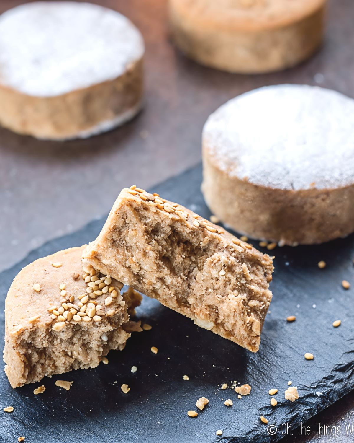 Crumbly Spanish Mantecados and Polvorones shortbread cookies topped with toasted sesame seeds and powdered sugar displayed on a dark slate board, served as a classic European Christmas dessert.