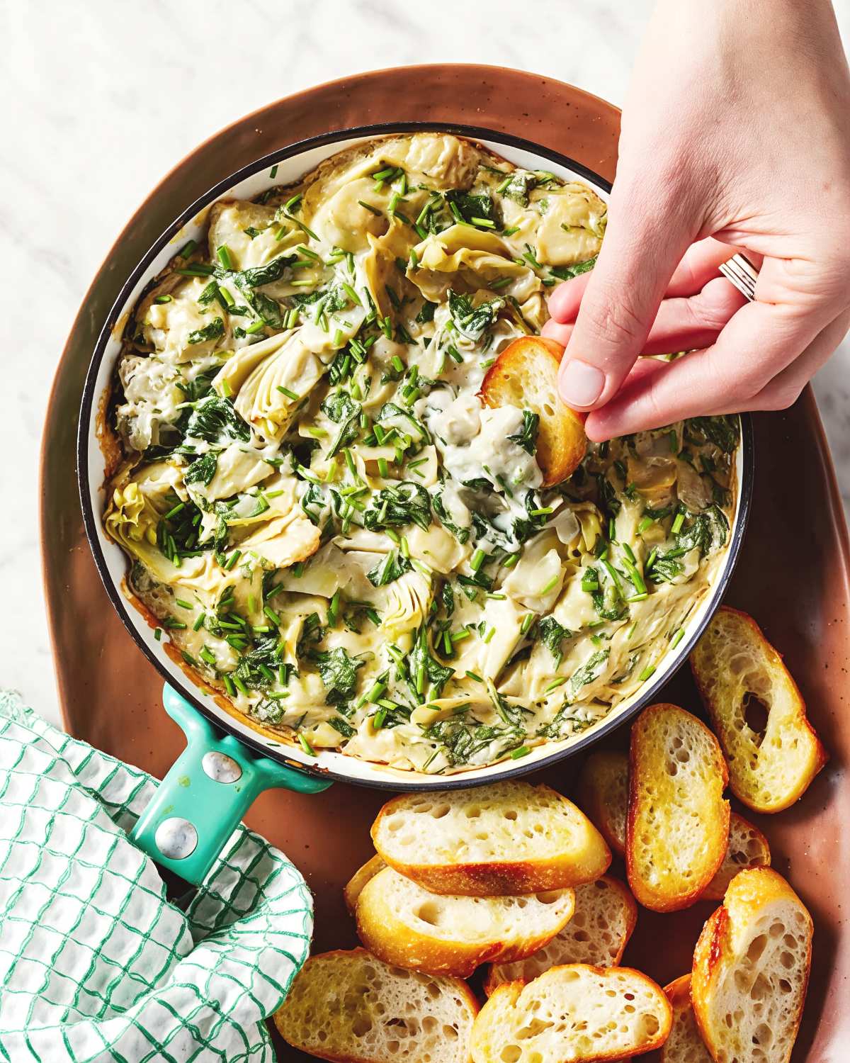 A hand dipping a toasted baguette slice into warm vegan spinach artichoke dip, served as one of the best vegan Thanksgiving appetizers.