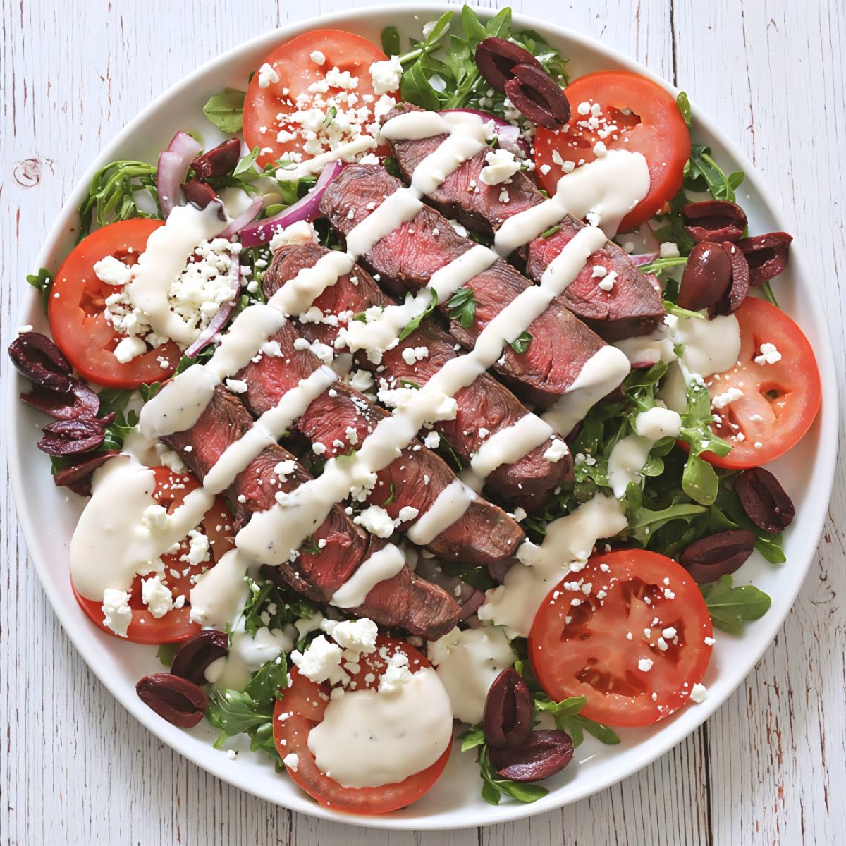 Top-down view of a fresh Mediterranean-style Steak Salad with sliced grilled steak, sliced tomatoes, red onions, feta cheese, and olives over leafy greens, featured as a delicious high protein low carb recipes dinner