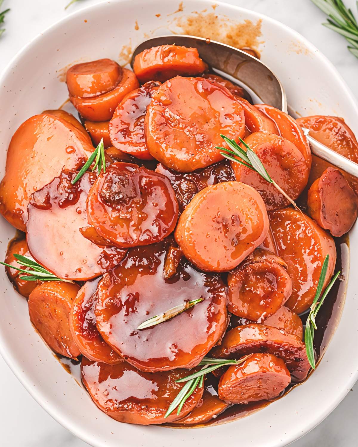 Stovetop candied sweet potatoes in a white serving dish coated with a thick brown sugar glaze and fresh rosemary as a Southern Thanksgiving side dish.
