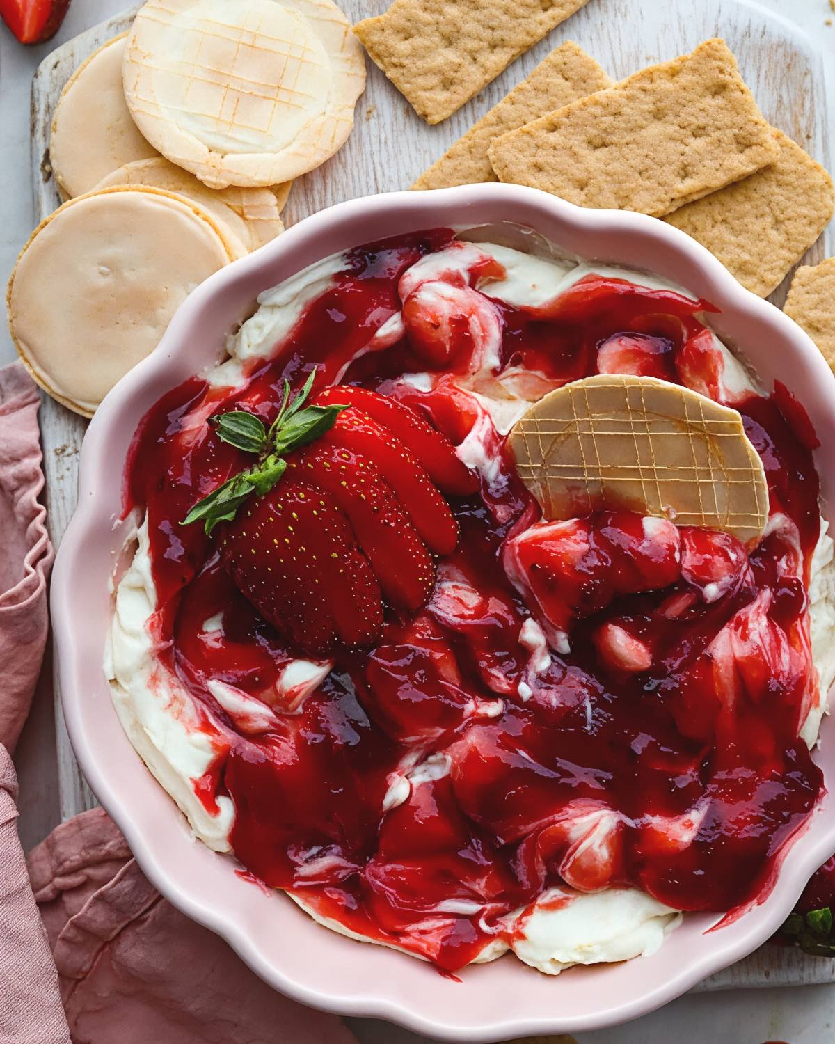 Creamy strawberry cheesecake dip topped with sweet strawberry sauce, fresh strawberry slices, graham crackers, and waffle cookies in a bowl, perfect as quick and easy last minute potluck ideas.