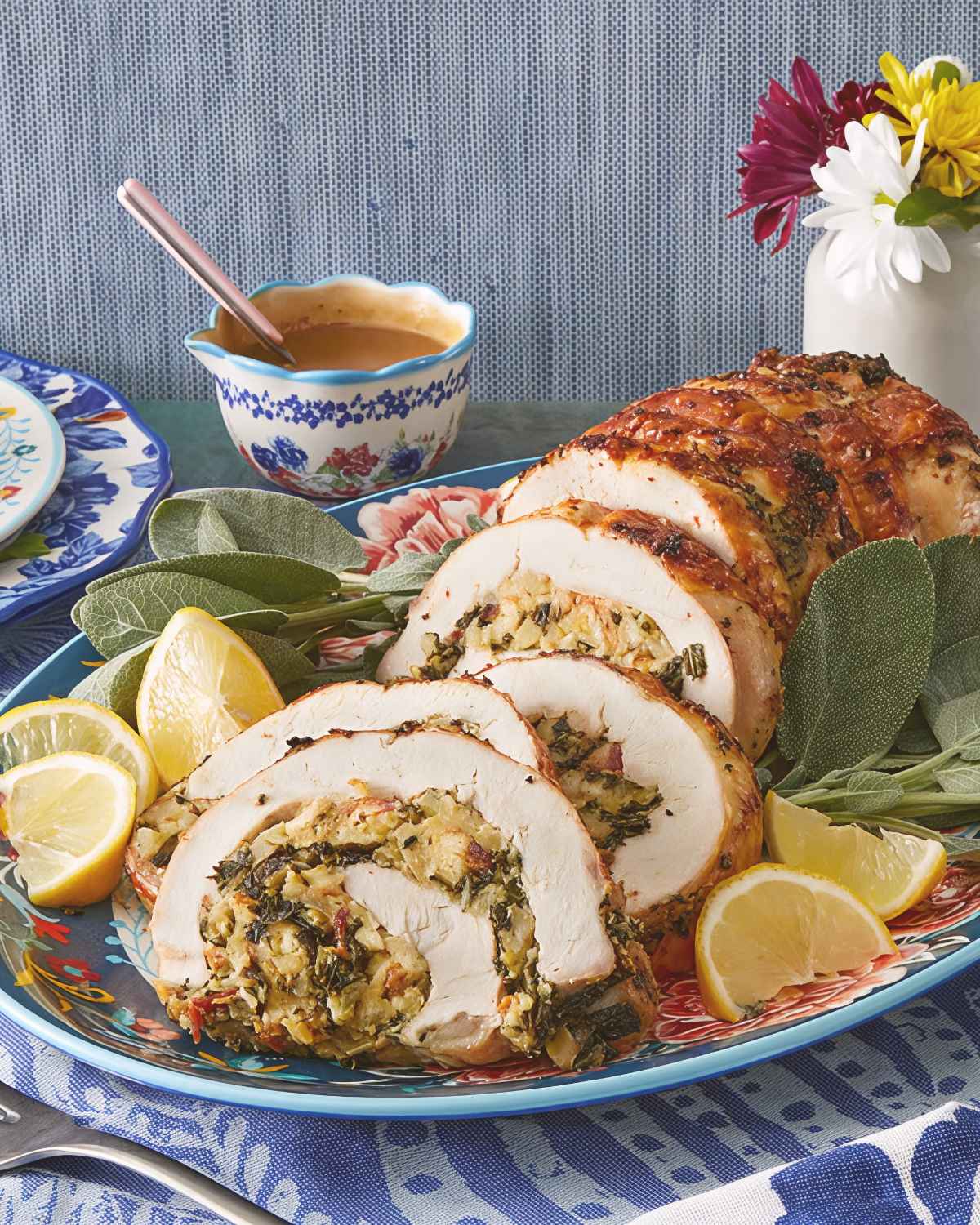 Sliced roasted turkey breast stuffed with herbs, spinach, and bread filling is arranged on a floral serving platter with fresh sage leaves and lemon wedges.