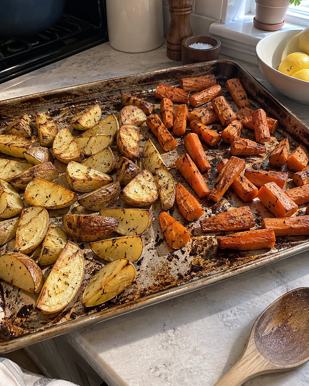 Carrot pieces with browned edges beside potatoes on a worn baking tray.