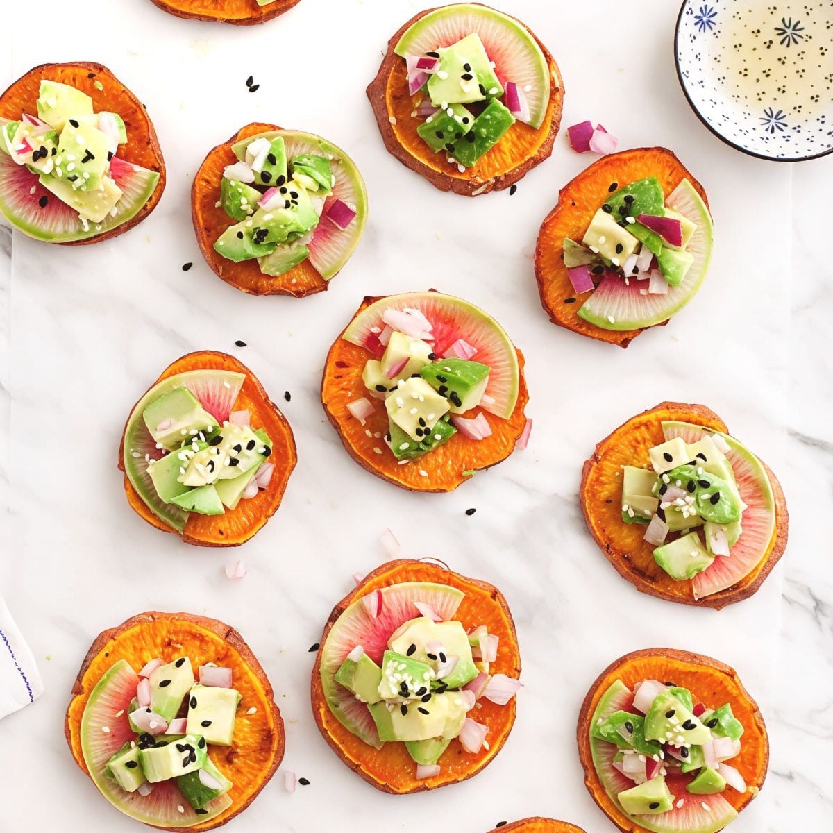 Roasted sweet potato appetizer bites topped with watermelon radish, avocado salsa, and sesame seeds arranged on a marble surface for Thanksgiving finger food ideas.