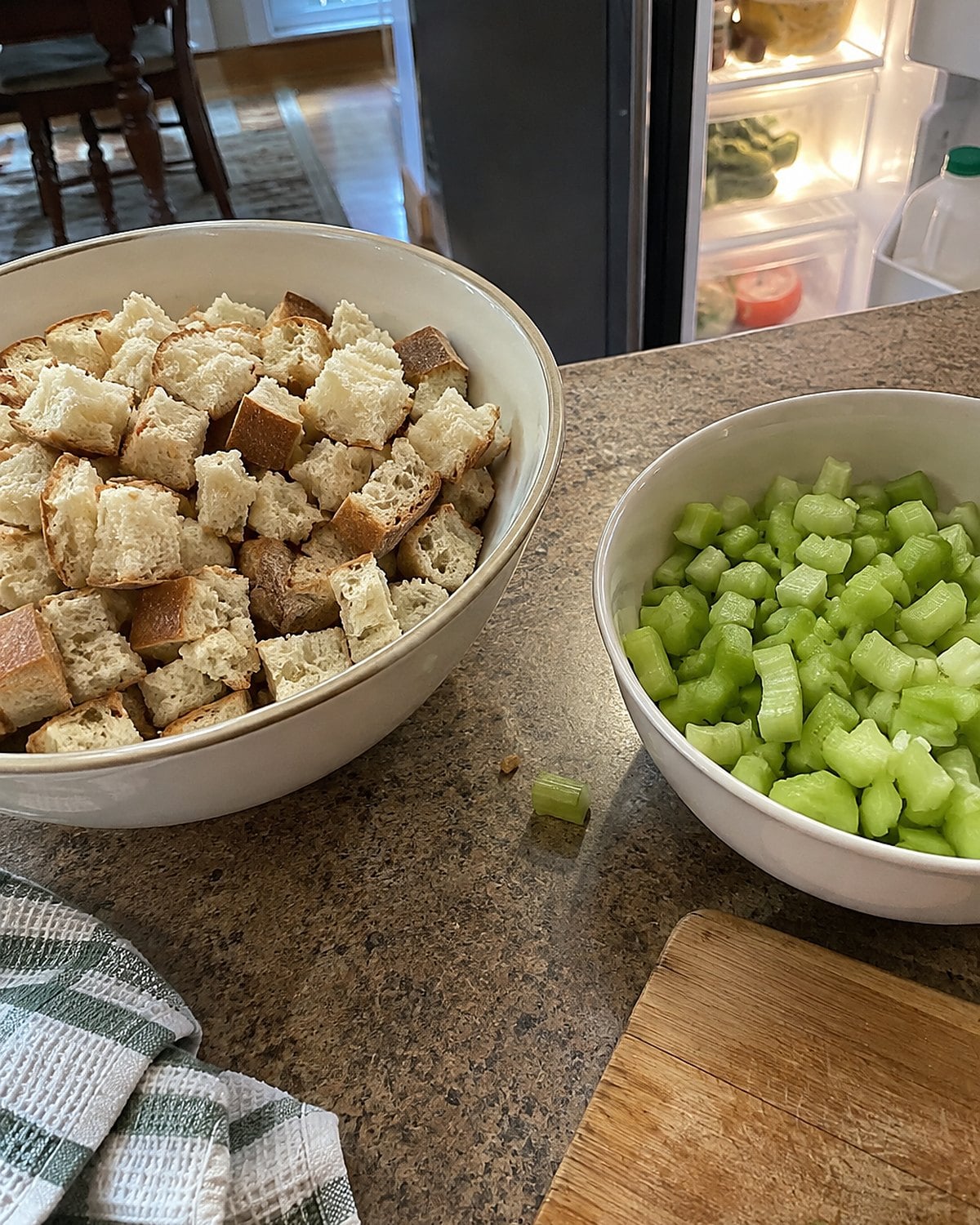 Separate bowls of dry bread cubes and chopped celery on a counter beside an open refrigerator.