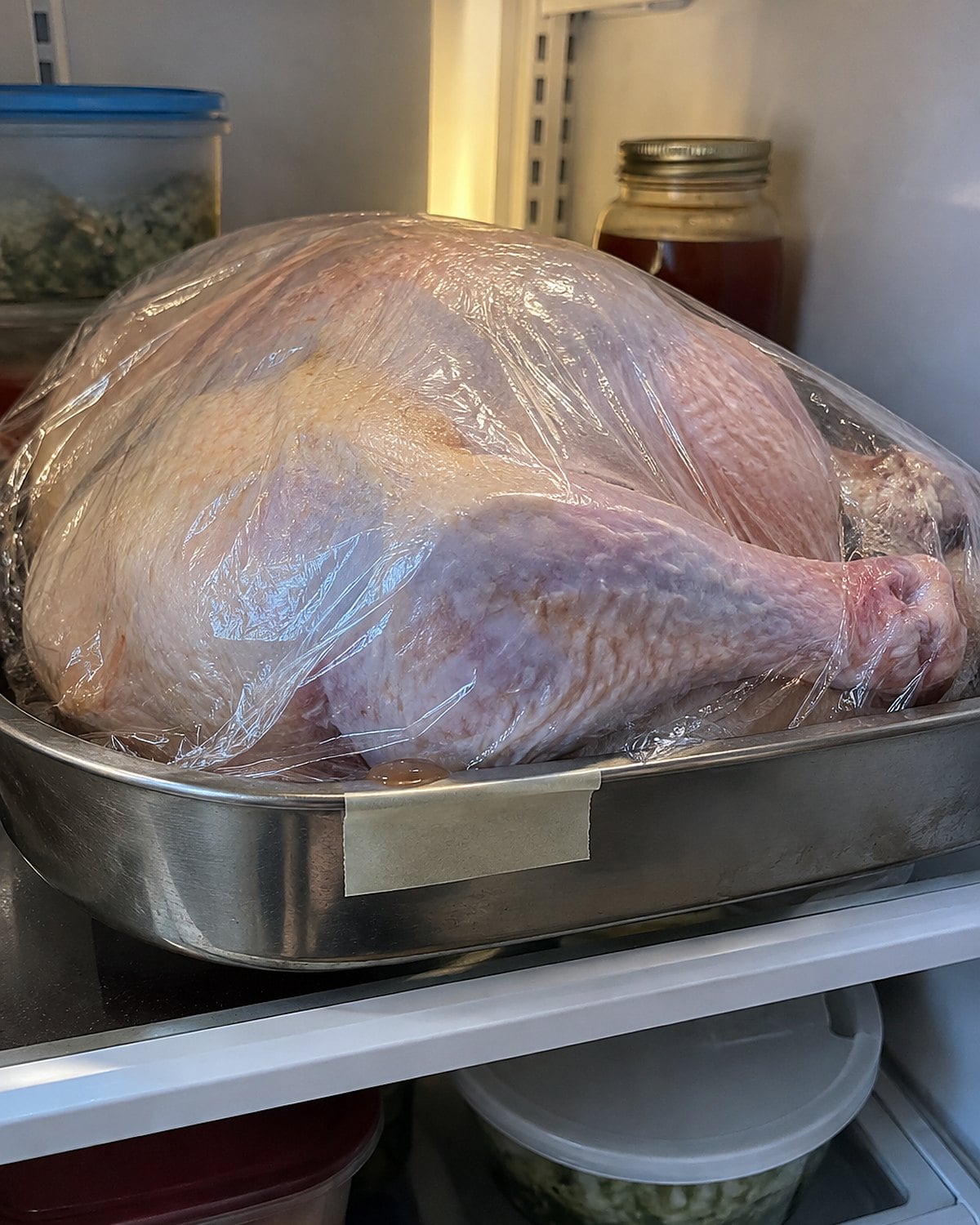 A wrapped turkey in a rimmed pan inside a refrigerator, with a handwritten date on tape attached to the pan.
