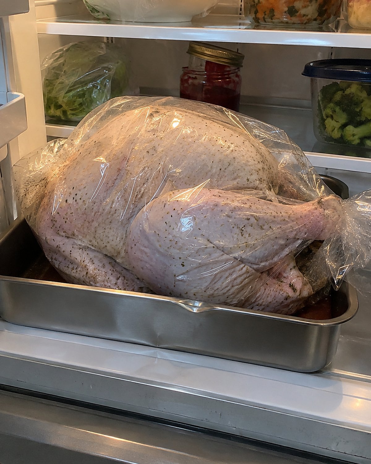 A wrapped turkey in a rimmed pan on the lowest refrigerator shelf, with a small drip caught inside the pan.