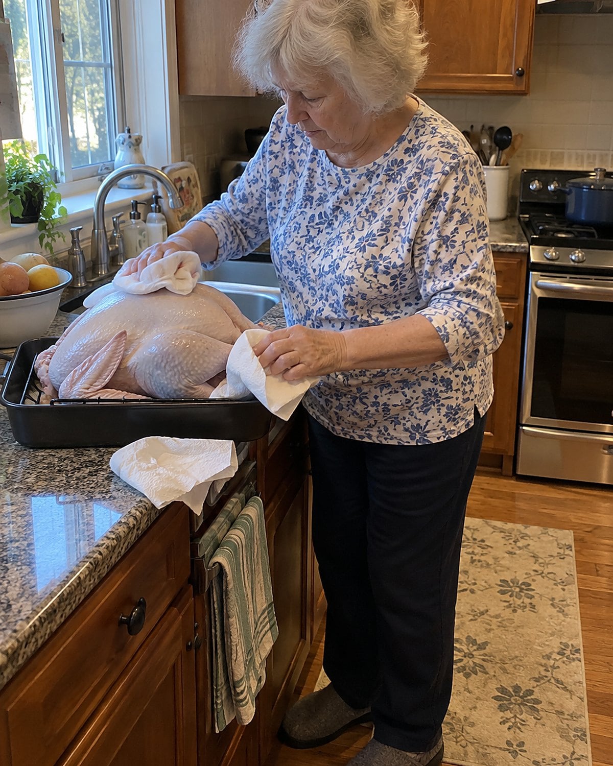 An older womanโs hands patting a turkey dry with paper towels over its roasting pan near the sink.