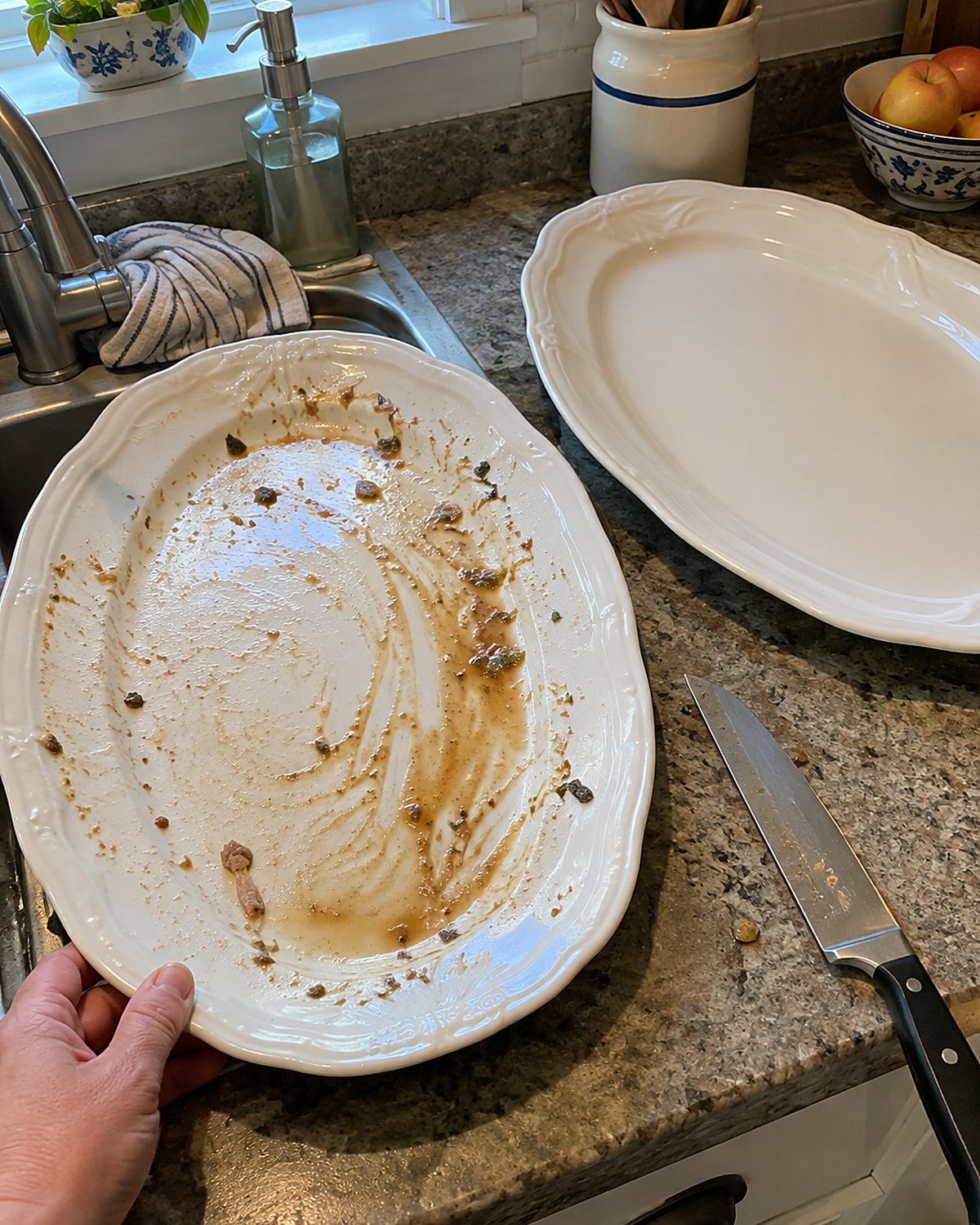 Two oval platters on a kitchen counter, an empty unwashed platter by the sink and a clean one beside a carving knife.
