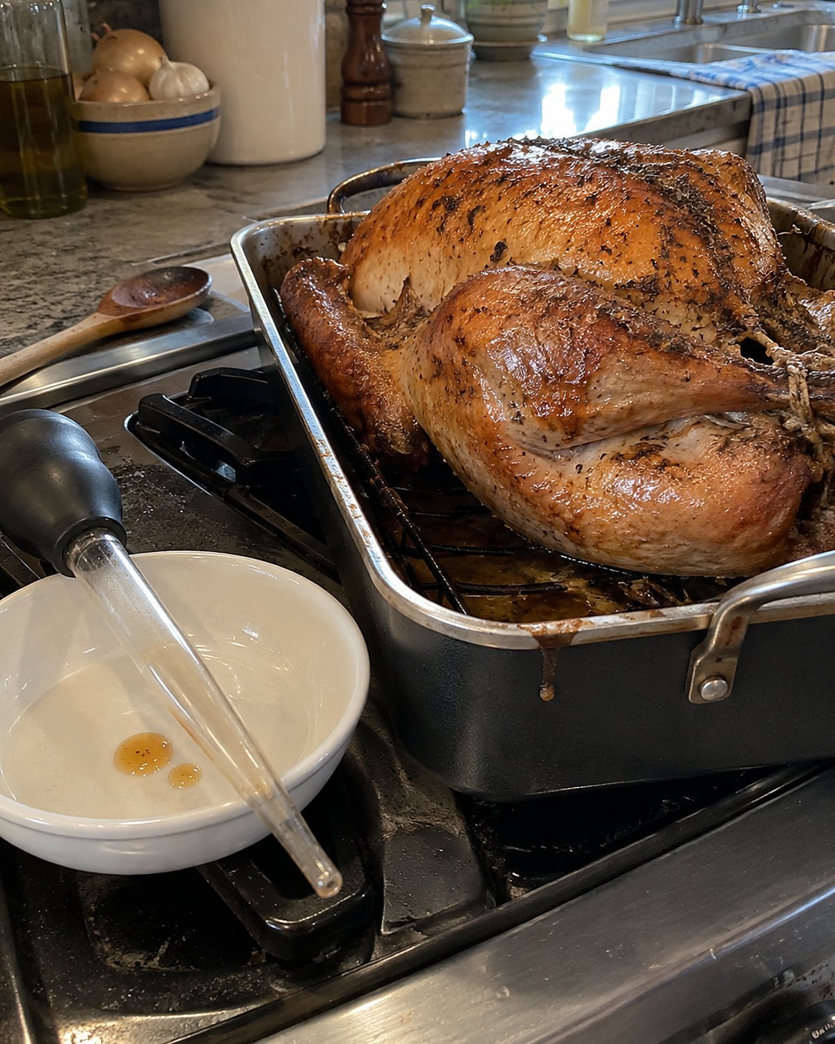 A turkey baster resting in a small dish beside a roasting pan on the stove.