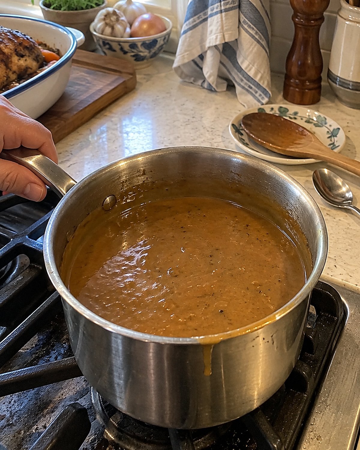 A saucepan of gravy on the stove with a cooking spoon resting on a saucer and a clean spoon beside the gravy boat.