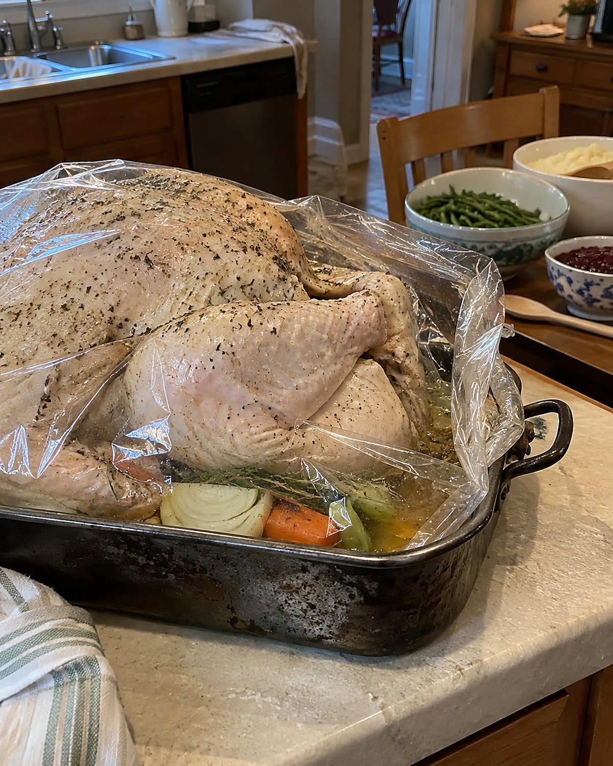 A roasting pan holding a wrapped turkey on a cleared counter, with serving bowls crowded on a nearby chair.