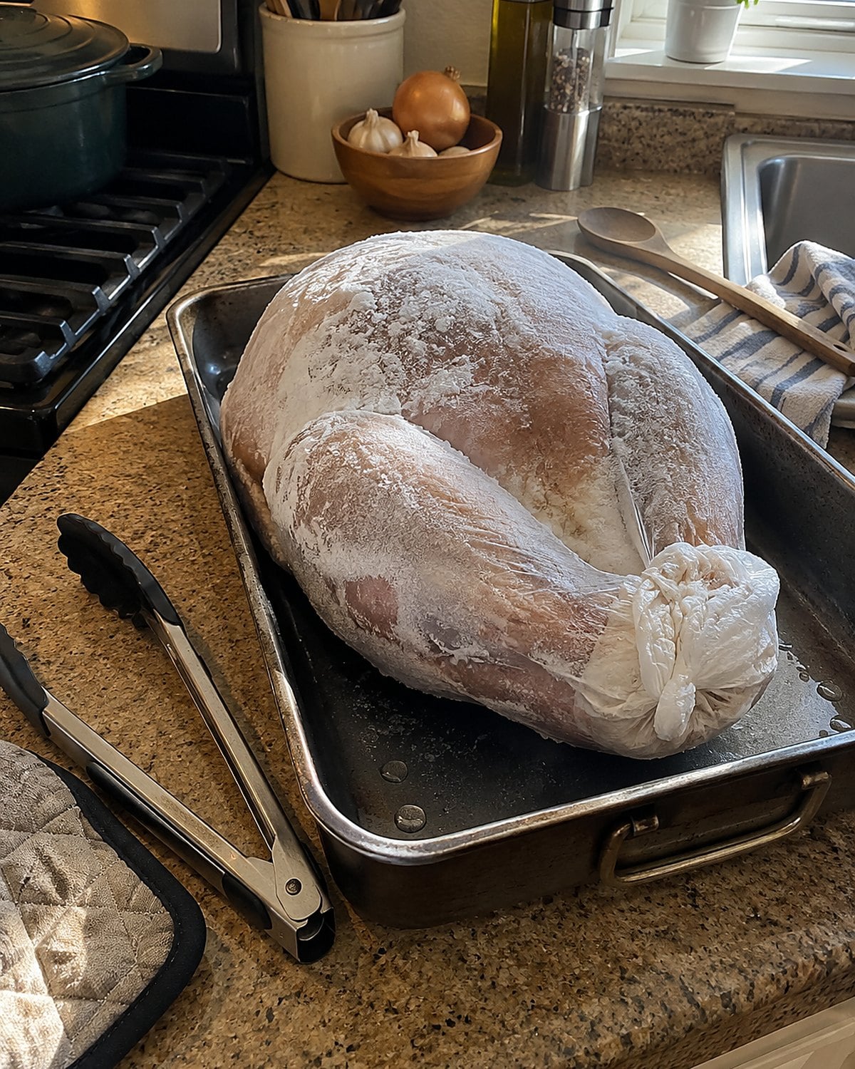 A frozen turkey in a roasting pan at the stove, with tongs and an oven mitt beside it.