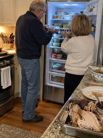 A couple in their sixties at the open fridge door the day after Thanksgiving, evening stove light on their hands as they shift foil-covered dishes across full shelves, nobody posing.